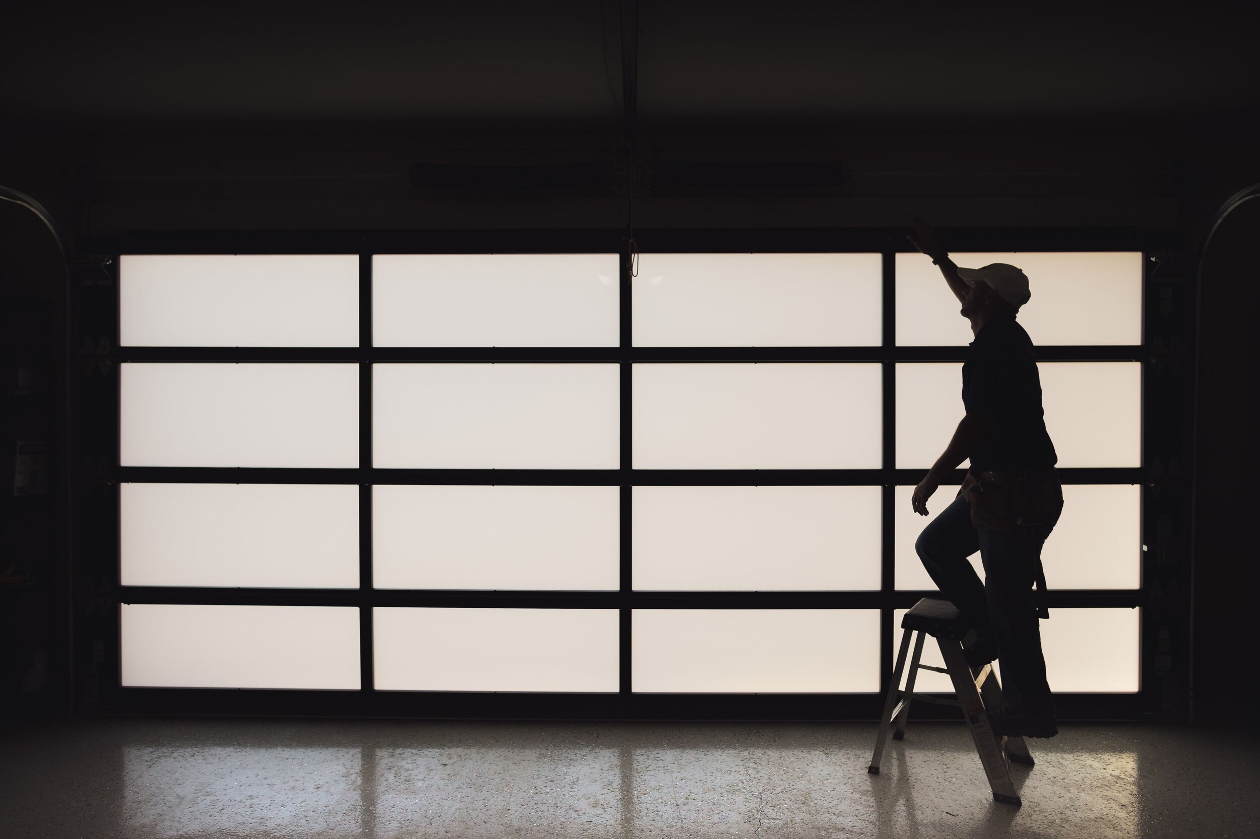 Silhouette of a worker on a ladder, reaching towards a garage door during home improvement or maintenance.