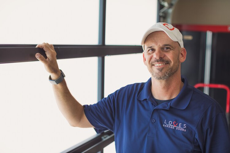 Man in a blue shirt and cap smiles while standing near large windows, showcasing garage door services.