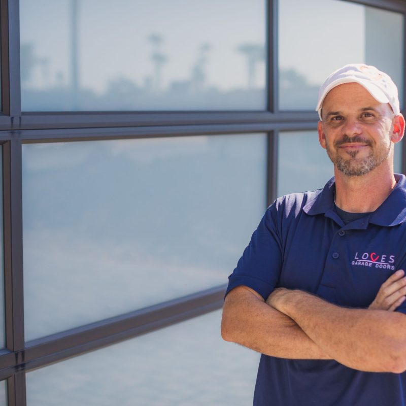 Confident man in a blue shirt with 'Loves Garage Doors' logo stands by a modern garage door.