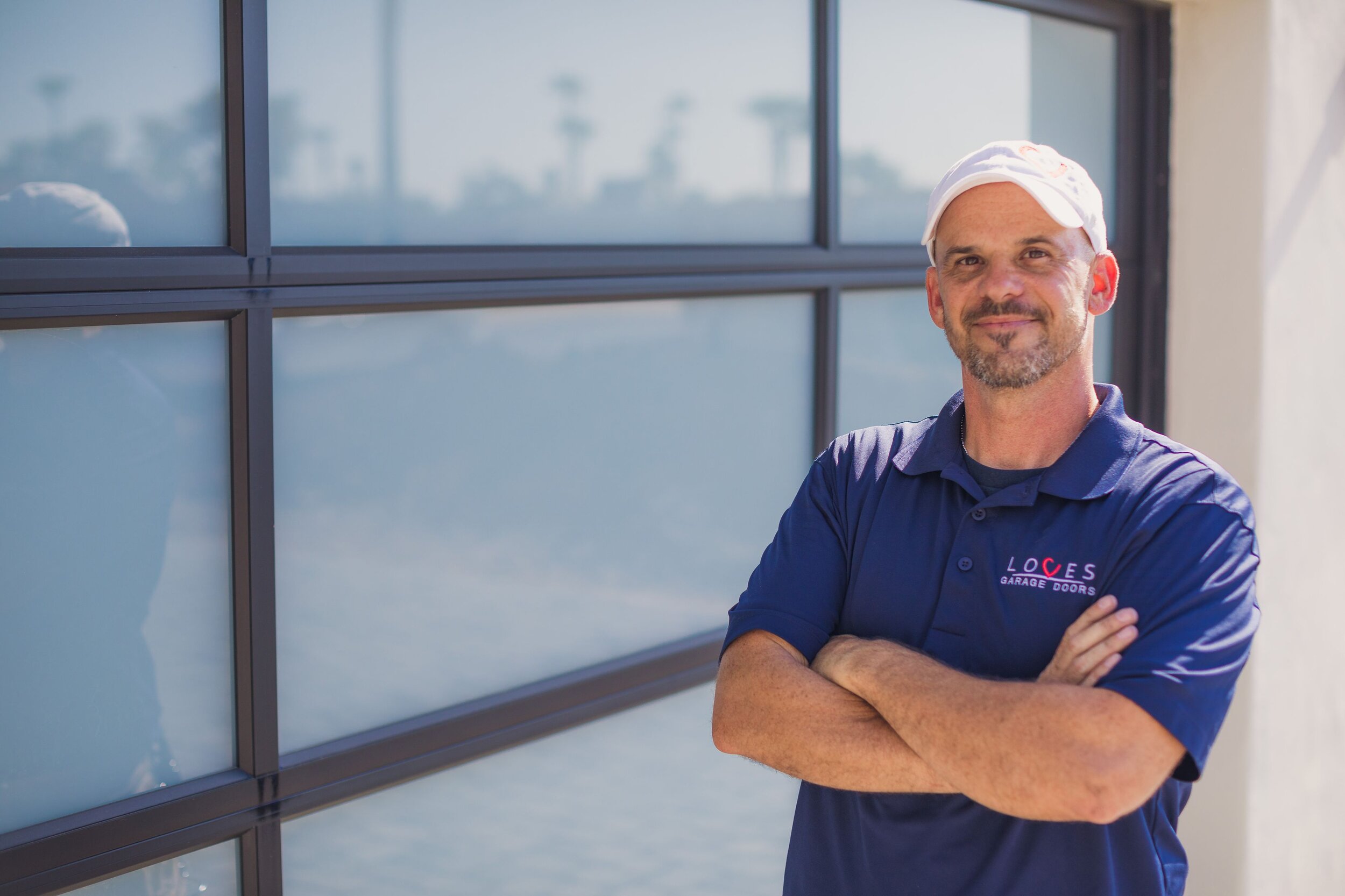 Confident man in a blue shirt with 'Loves Garage Doors' logo stands by a modern garage door.