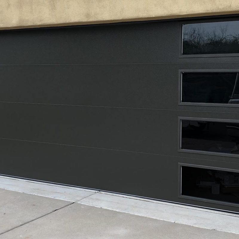 Modern dark garage door with three glass panels, set against a beige wall and surrounded by greenery.