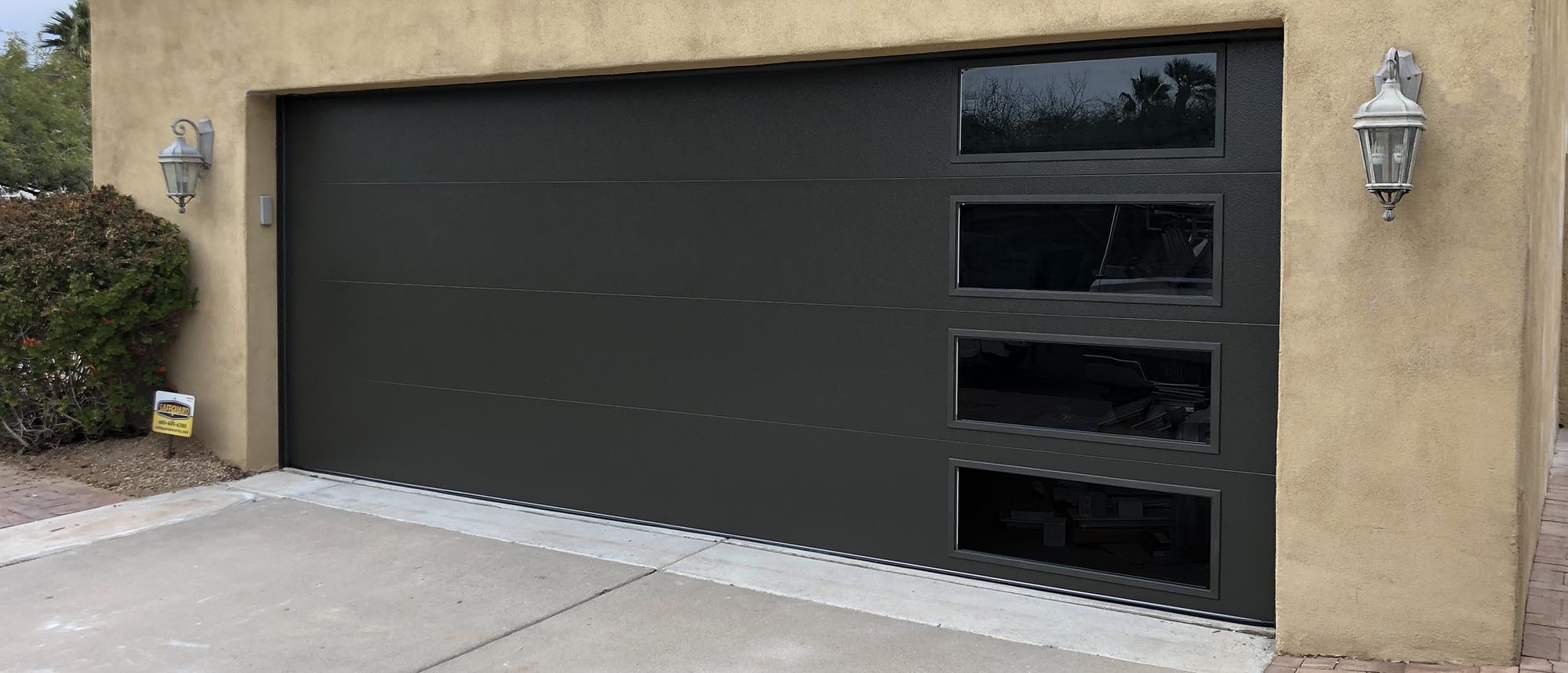 Modern dark garage door with three glass panels, set against a beige wall and surrounded by greenery.