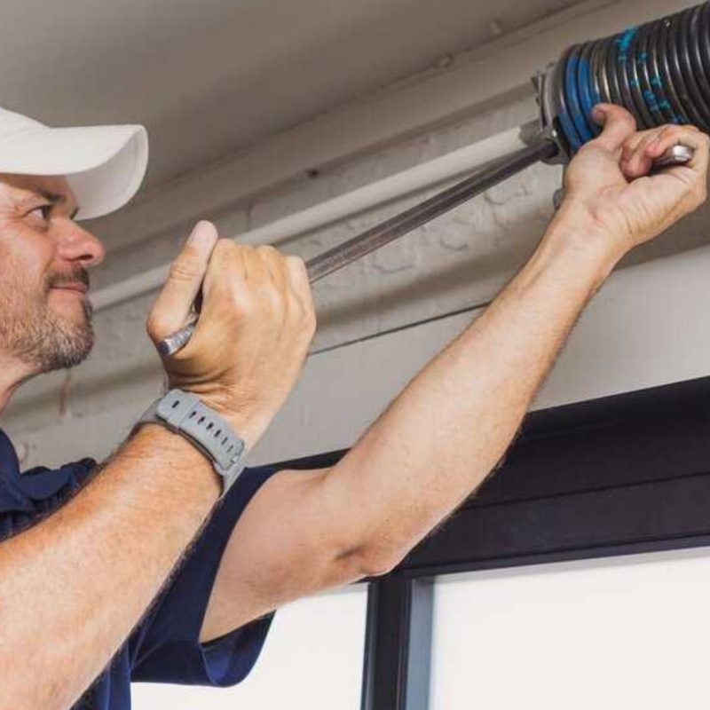 A technician wearing a white cap and blue shirt uses a winding bar to adjust a garage door torsion spring.
