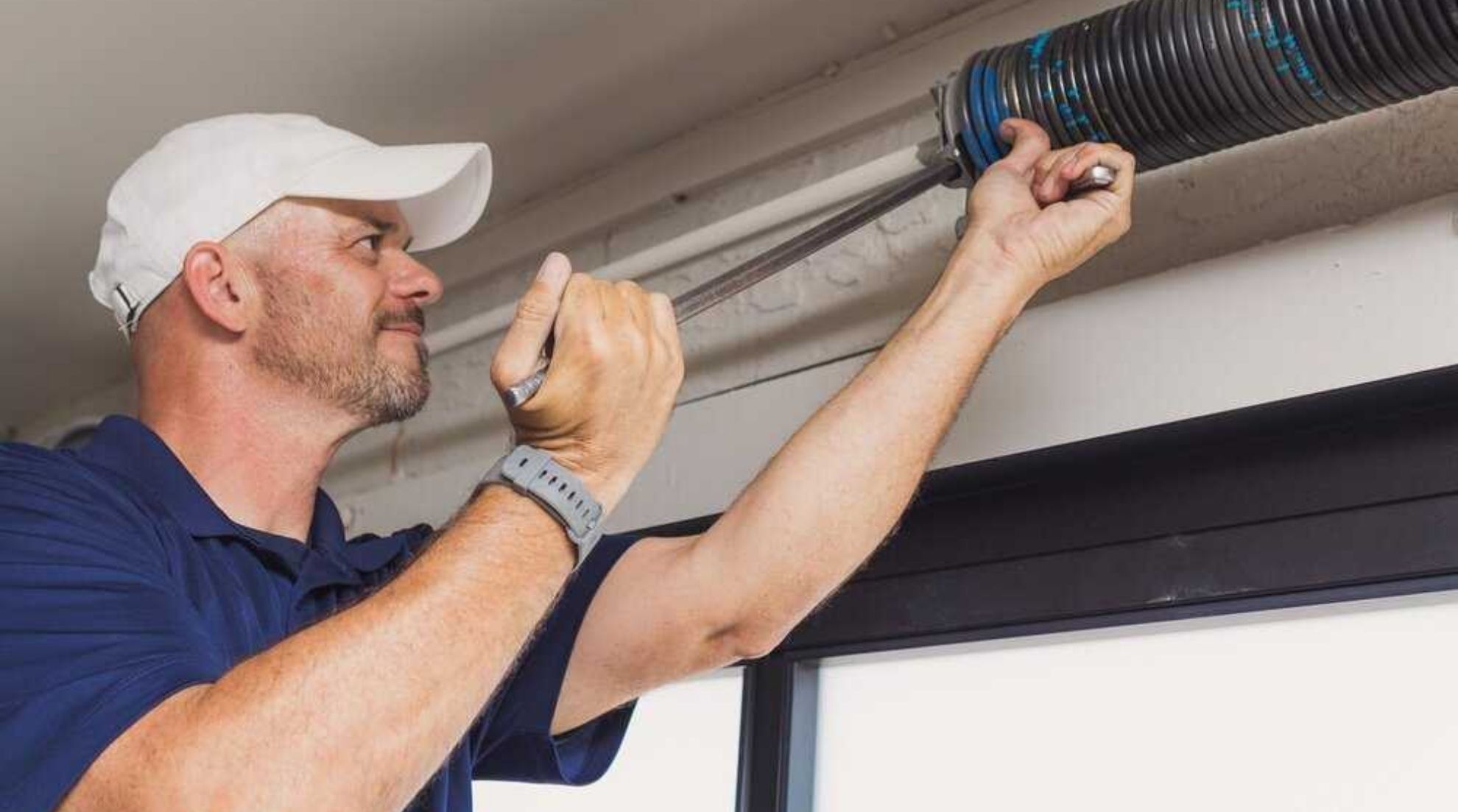 A technician wearing a white cap and blue shirt uses a winding bar to adjust a garage door torsion spring.