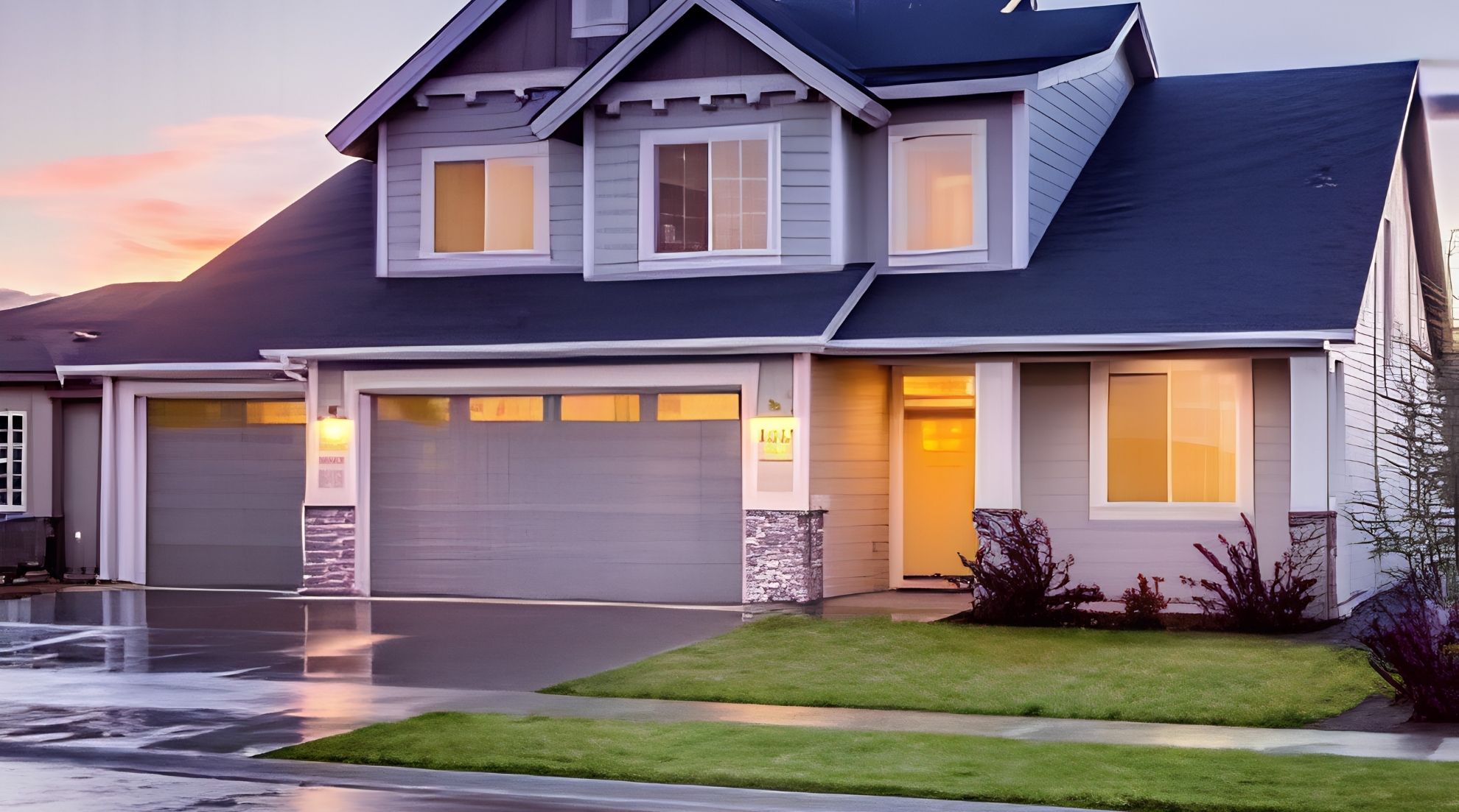 Modern two-story home at dusk with a closed double-car garage door illuminated by warm exterior lighting.