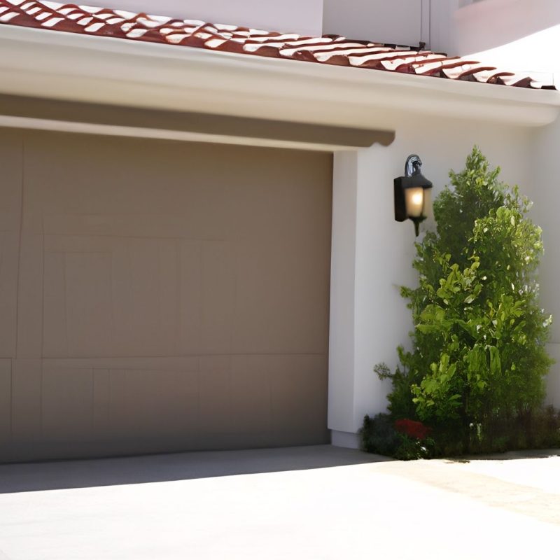 Modern beige garage door on a white house with a green shrub, potted plant, and outdoor wall lanterns.
