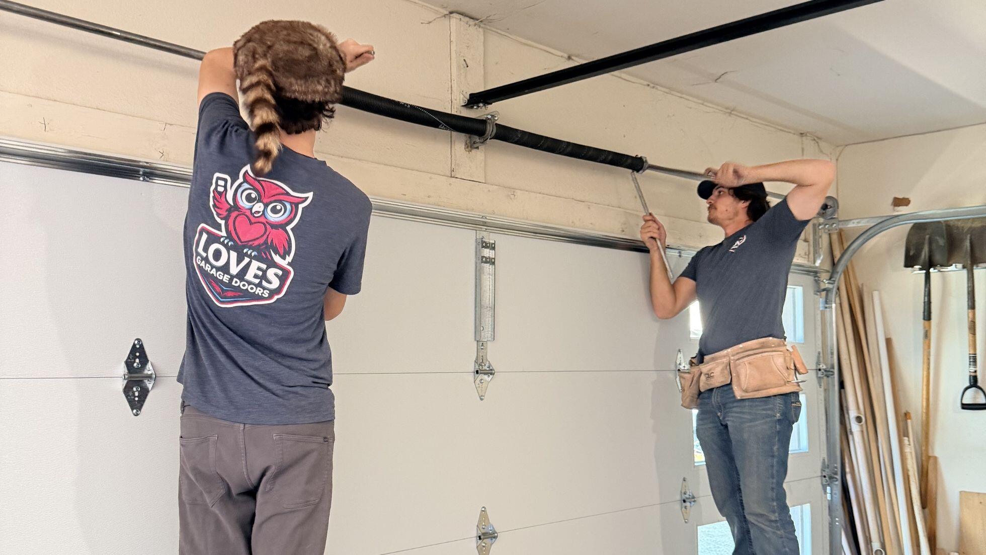 Two men repairing a garage door, focusing on spring installation with tools and equipment in a home garage.