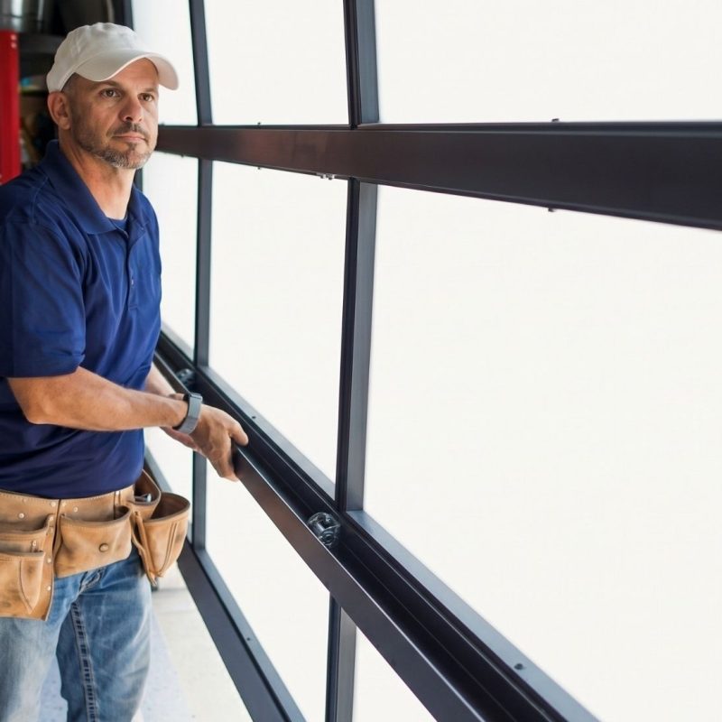 Man in a blue polo shirt and tool belt installing a garage door in a bright workspace filled with tools and supplies.